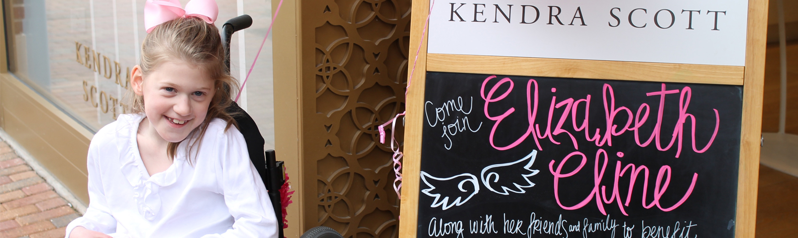 Little girl in a wheelchair next to a sign saying there is an event benefitting the Angelman Syndrome Foundation