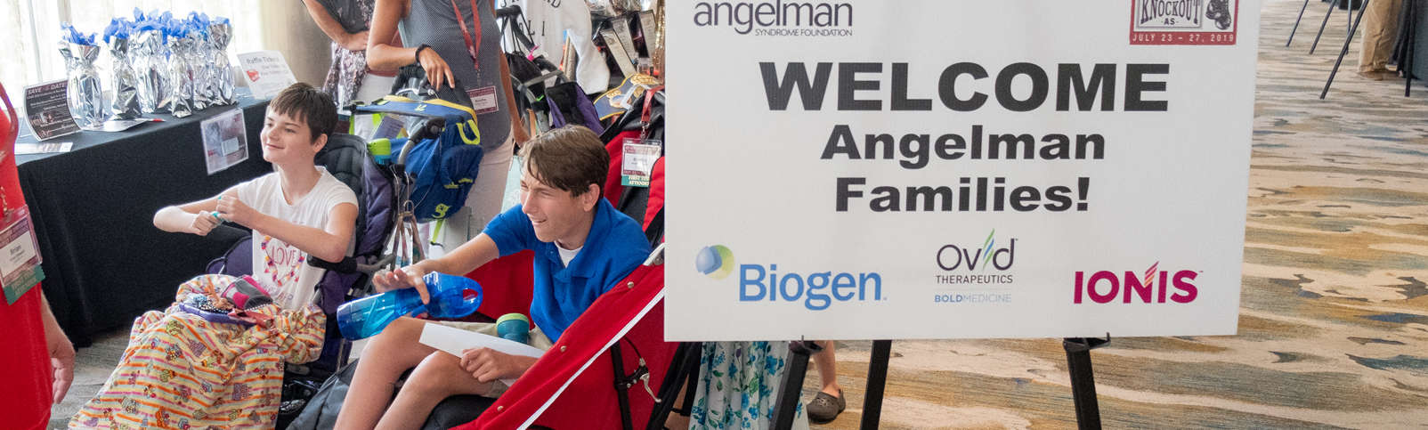 Two individuals with AS next to a sign that welcomes Angelman families